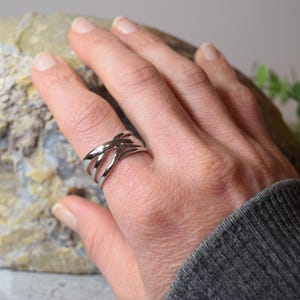 Peut inclure: Une bague en métal argenté poli présentant un design multi-anneaux avec des brins métalliques qui se croisent et se chevauchent autour du doigt. La bague possède une surface lisse et réfléchissante ainsi qu'une structure ajourée et large.