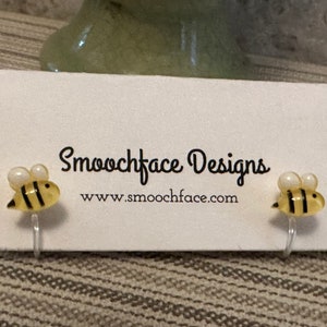 May include: A pair of yellow and black bee stud earrings with white pearl accents. The earrings are on a white card with the text "Smoochface Designs" and "www.smoochface.com".