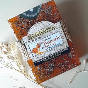 Puede incluir: Una pastilla rectangular de limpiador facial con un diseño moteado naranja y marrón. La etiqueta dice "Organic Tumeric & Honey with Blue Poppy" y "Earthen Urban Essentials 100% Pure & Natural". La pastilla está envuelta con una fibra natural.