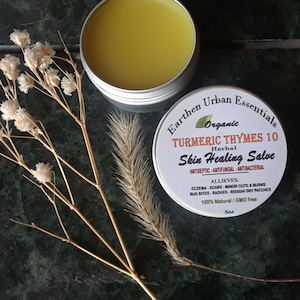 May include: A round silver metal tin containing yellow herbal salve, with a lid labeled Earthen Urban Essentials Organic Turmeric Thymes 10 Herbal Skin Healing Salve, Antiseptic-Antifungal-Antibacterial, Allieves: Eczema-Scars-Minor Cuts & Burns, .5oz.