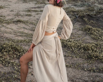 May include: A woman wearing a white, long-sleeved, wrap top and a matching white maxi skirt. She is wearing a pink flower crown and is standing on a sandy beach with green vines.