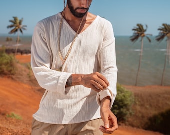 Puede incluir: Un hombre con una camisa blanca de manga larga con cuello en V, pantalones beige y un fedora blanco con una banda de cuentas. Está de pie en un camino de tierra roja con palmeras en el fondo.