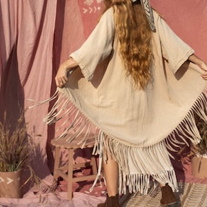 以下が含まれることがあります： ピンクの背景の前に立っている女性。女性は、薄い茶色のフリンジ付きのキモノを着て、茶色の帽子をかぶっています。帽子には白と茶色の模様のバンドが付いています。