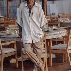Puede incluir: Una camisa de botones de color beige claro de corte holgado con mangas cortas y anchas y cuello, combinada con pantalones de color tostado de corte suelto que presentan un detalle de dobladillo con vuelta en los tobillos. La camisa tiene botones.