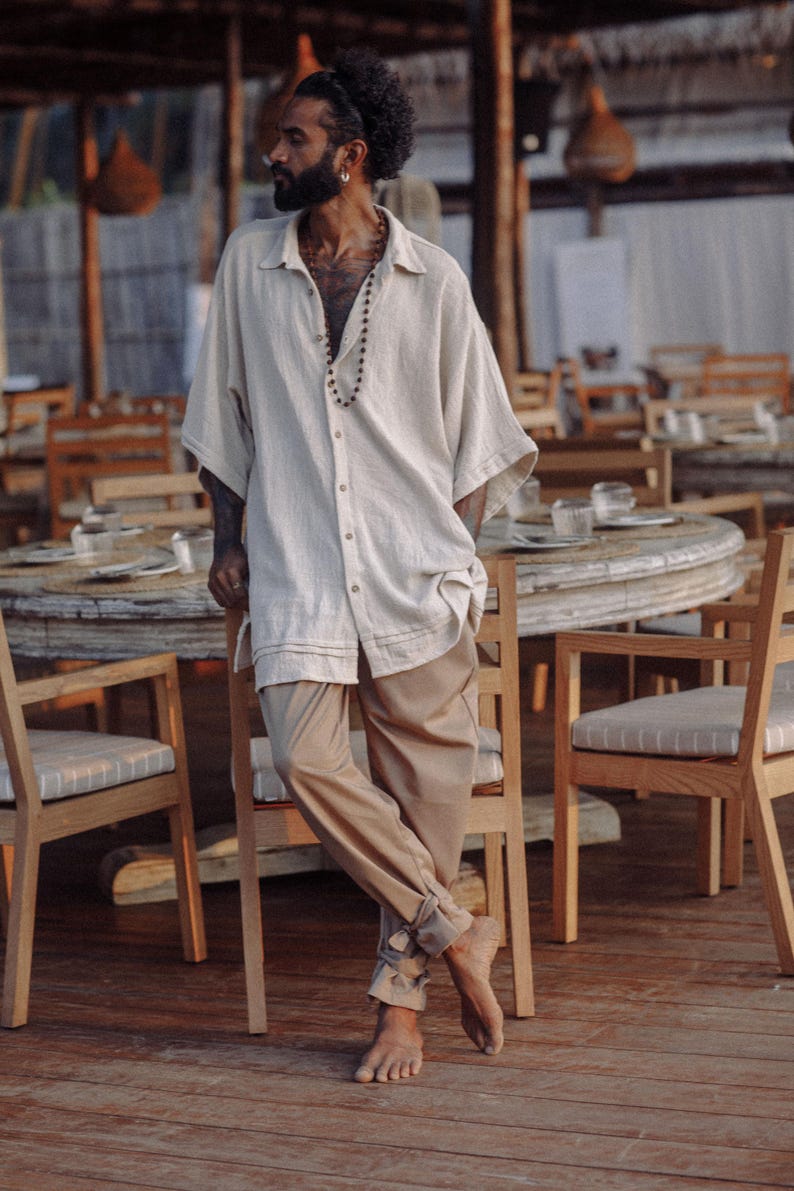 Puede incluir: Una camisa de botones de color beige claro de corte holgado con mangas cortas y anchas y cuello, combinada con pantalones de color tostado de corte suelto que presentan un detalle de dobladillo con vuelta en los tobillos. La camisa tiene botones.