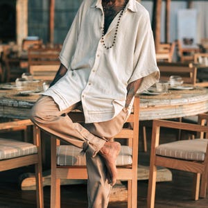 Puede incluir: Una camisa de botones de color blanco roto, de manga corta, con cuello y corte holgado, combinada con pantalones de color tostado que presentan detalles de lazos en los tobillos. La camisa tiene una textura visible y un collar de cuentas.