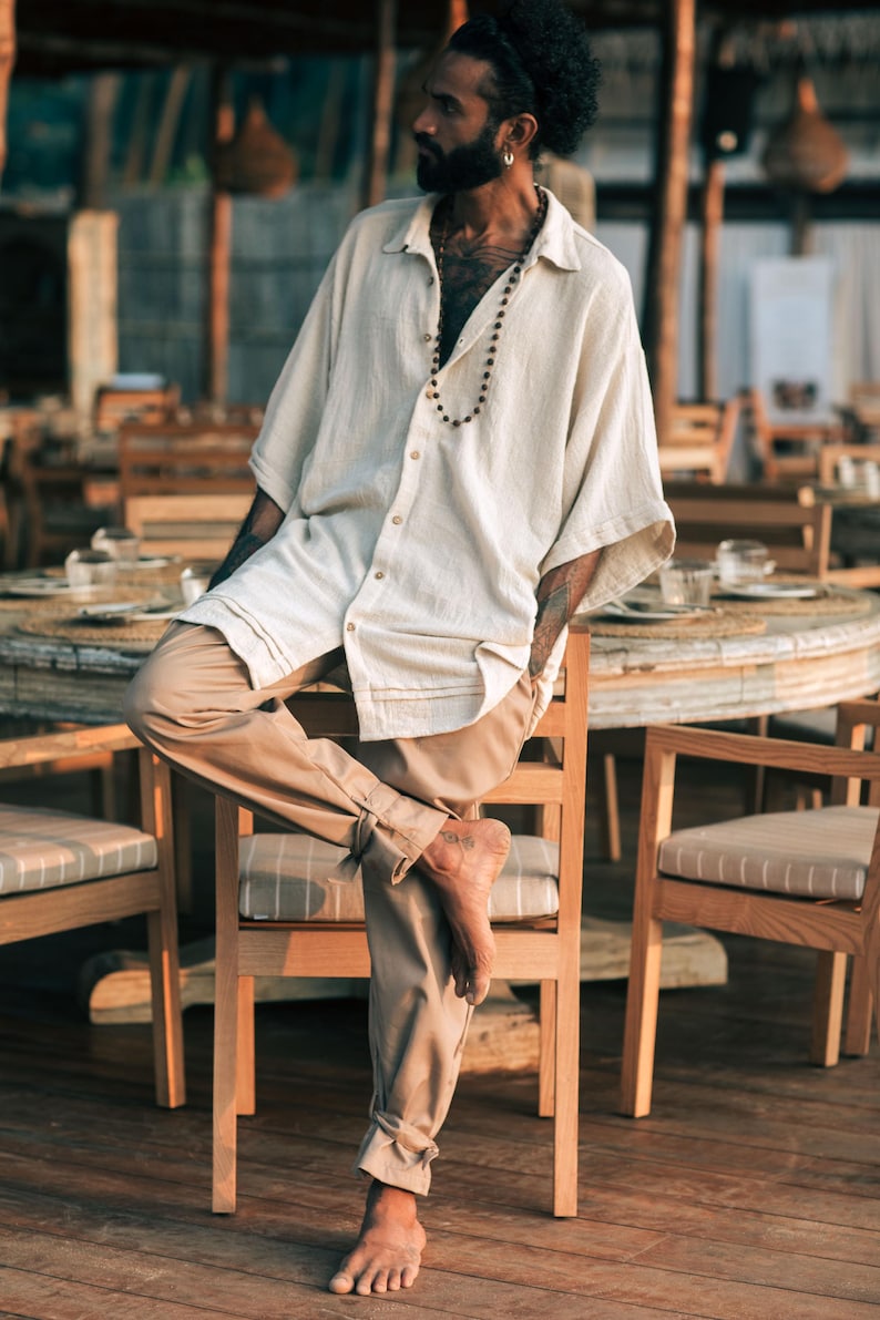 Puede incluir: Una camisa de botones de color blanco roto, de manga corta, con cuello y corte holgado, combinada con pantalones de color tostado que presentan detalles de lazos en los tobillos. La camisa tiene una textura visible y un collar de cuentas.