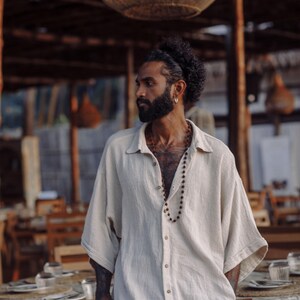 Puede incluir: Una camisa beige de corte oversize con botones, tejido texturizado, cuello cl&aacute;sico y mangas anchas hasta el codo. La prenda cuenta con cierre frontal de botones y un dobladillo recto, combinada con un collar largo de peque&ntilde;as cuentas de color marr&oacute;n.