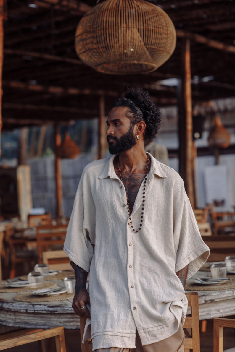 Puede incluir: Una camisa beige de corte oversize con botones, tejido texturizado, cuello cl&aacute;sico y mangas anchas hasta el codo. La prenda cuenta con cierre frontal de botones y un dobladillo recto, combinada con un collar largo de peque&ntilde;as cuentas de color marr&oacute;n.