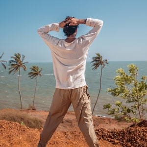 May include: A person wearing a white long-sleeved shirt and beige pants stands on a red dirt cliff overlooking the ocean. The person has their arms raised above their head and is looking out at the ocean. There are palm trees in the background.