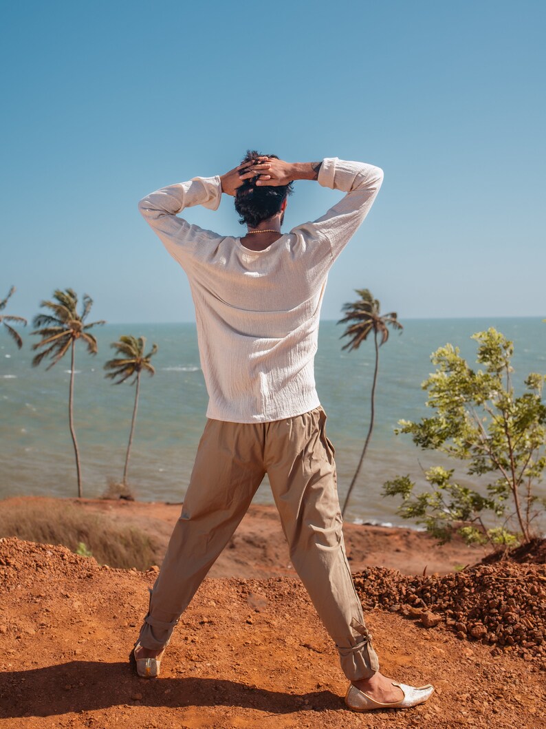 May include: A person wearing a white long-sleeved shirt and beige pants stands on a red dirt cliff overlooking the ocean. The person has their arms raised above their head and is looking out at the ocean. There are palm trees in the background.
