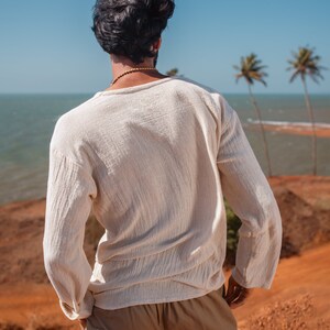 May include: A person wearing a white long-sleeved shirt and khaki pants stands on a red dirt hill overlooking a blue ocean and palm trees.