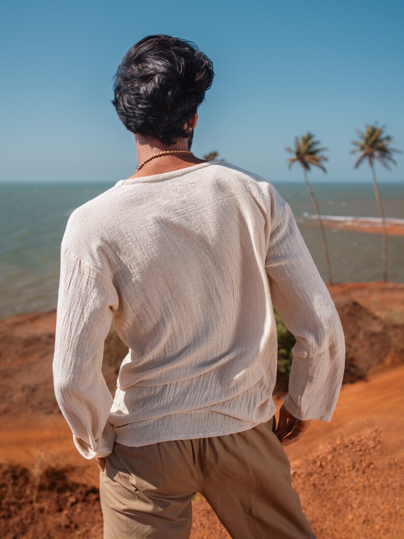 May include: A person wearing a white long-sleeved shirt and khaki pants stands on a red dirt hill overlooking a blue ocean and palm trees.