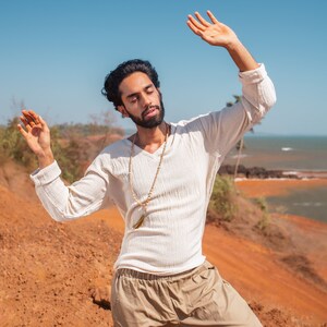 May include: A man wearing a white long-sleeved shirt with a brown beaded necklace and beige pants, standing with his arms raised in a relaxed pose against a backdrop of a red dirt hill and blue sky.