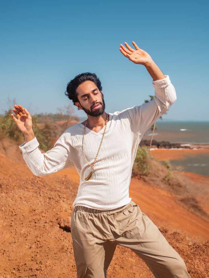 May include: A man wearing a white long-sleeved shirt with a brown beaded necklace and beige pants, standing with his arms raised in a relaxed pose against a backdrop of a red dirt hill and blue sky.