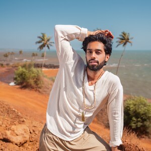 May include: A man wearing a white long-sleeved shirt with a beaded necklace and a tassel pendant. He is wearing khaki pants and has his hand on his head. He is standing in front of a red dirt path with a blue sky and ocean in the background.