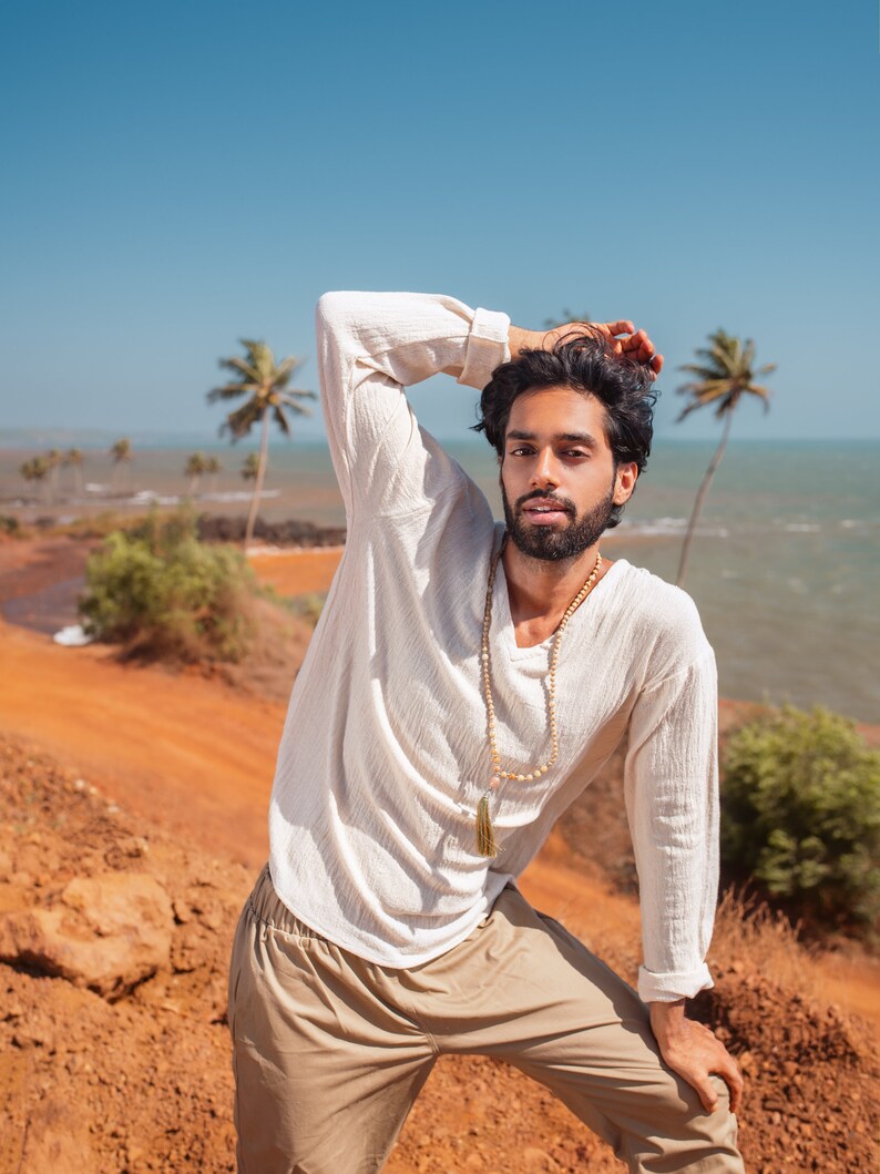 May include: A man wearing a white long-sleeved shirt with a beaded necklace and a tassel pendant. He is wearing khaki pants and has his hand on his head. He is standing in front of a red dirt path with a blue sky and ocean in the background.