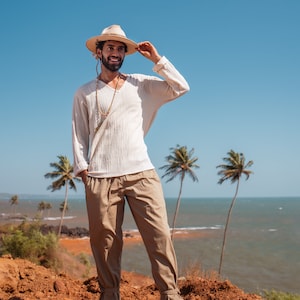 May include: A man wearing a white shirt, beige pants, and a straw hat with a beaded necklace. He is standing on a red dirt cliff overlooking the ocean with palm trees in the background.