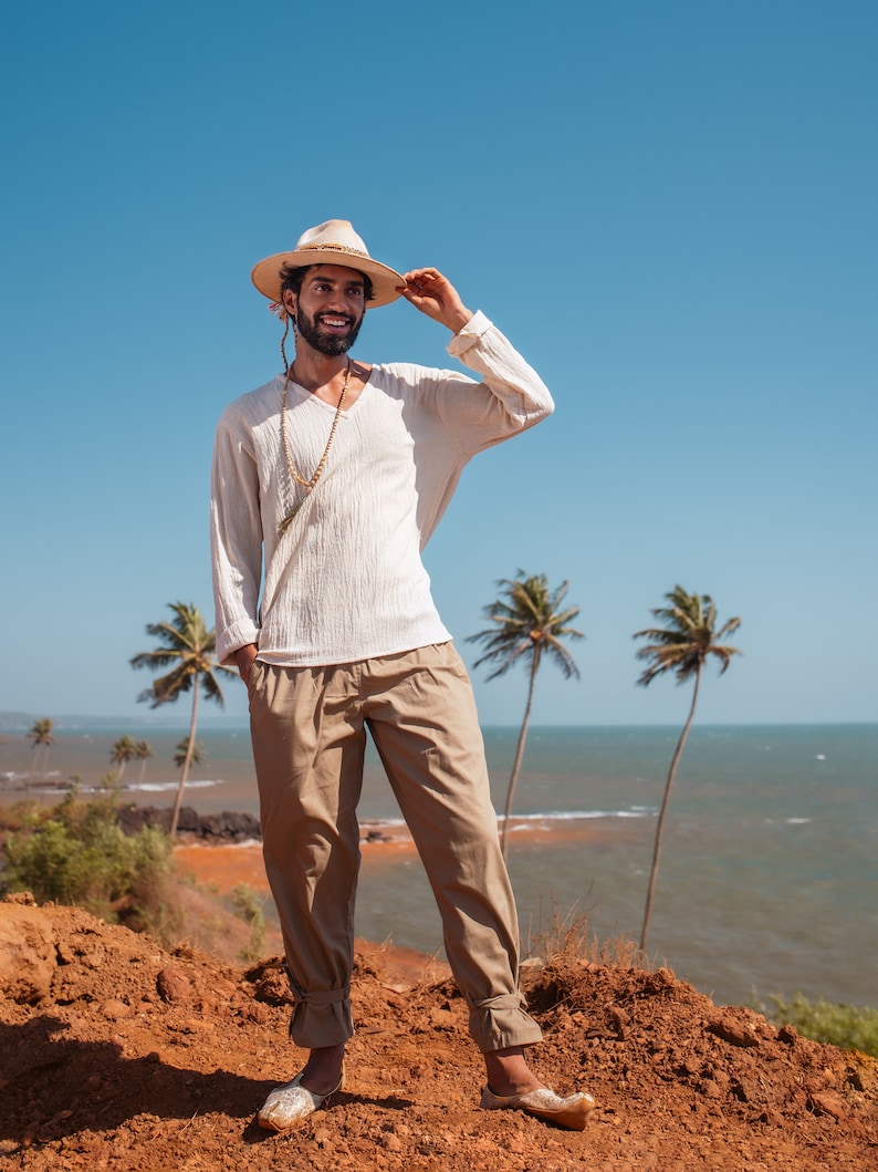 May include: A man wearing a white shirt, beige pants, and a straw hat with a beaded necklace. He is standing on a red dirt cliff overlooking the ocean with palm trees in the background.