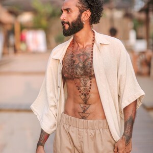 Puede incluir: Una camisa beige de textura de lino, desabotonada y de manga corta, combinada con pantalones de color tostado. El conjunto se complementa con un collar largo de cuentas esf&eacute;ricas de color marr&oacute;n oscuro y una diadema negra en el pelo rizado.