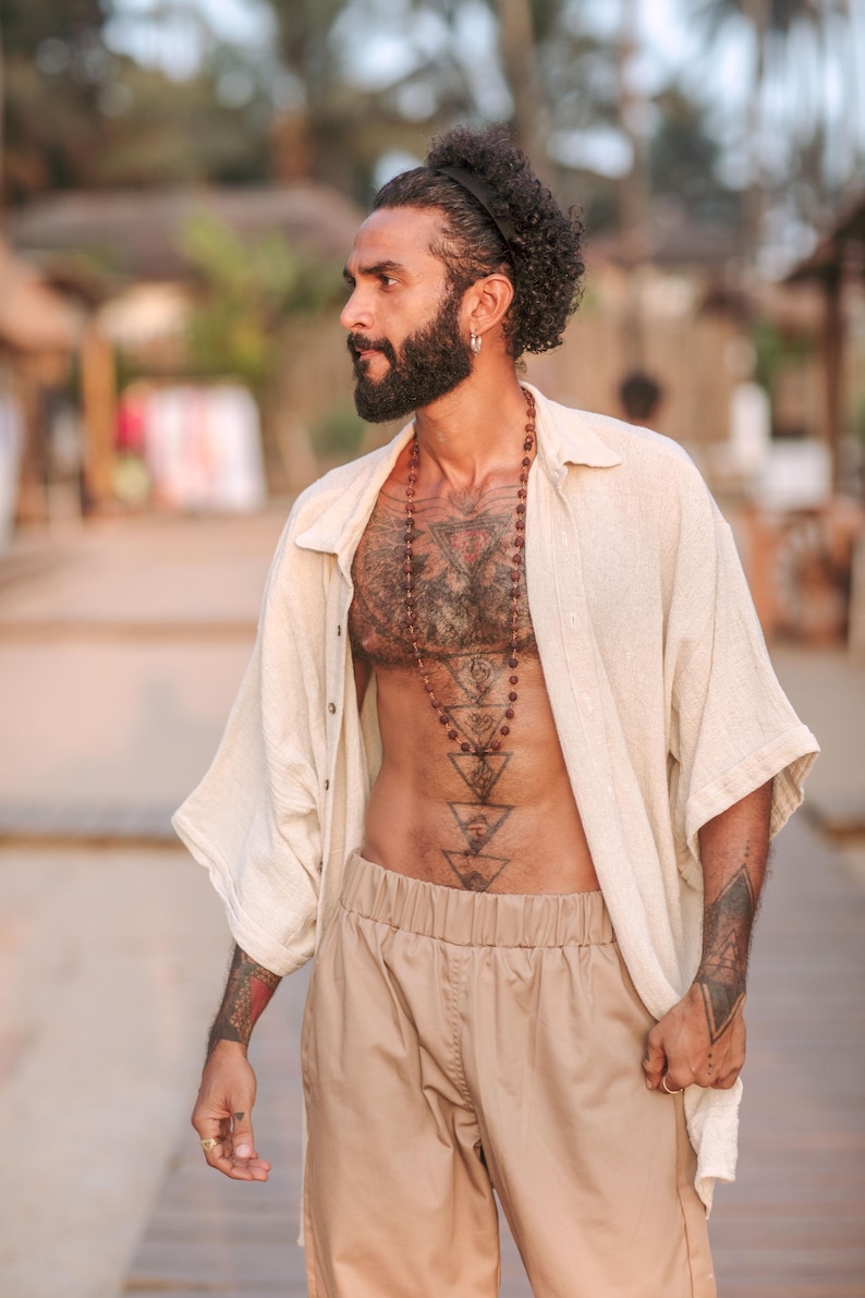 Puede incluir: Una camisa beige de textura de lino, desabotonada y de manga corta, combinada con pantalones de color tostado. El conjunto se complementa con un collar largo de cuentas esf&eacute;ricas de color marr&oacute;n oscuro y una diadema negra en el pelo rizado.