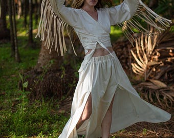 May include: Cream-colored wrap top with long sleeves and fringe detail, paired with a matching skirt featuring high slits. The outfit is worn outdoors in a forest setting, with the model wearing ankle boots.