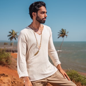 May include: A man wearing a white long-sleeved shirt with a beaded necklace and a gold tassel. He is standing on a red dirt path with a blue ocean and palm trees in the background.