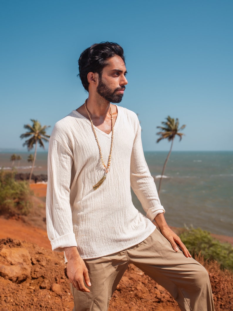 May include: A man wearing a white long-sleeved shirt with a beaded necklace and a gold tassel. He is standing on a red dirt path with a blue ocean and palm trees in the background.