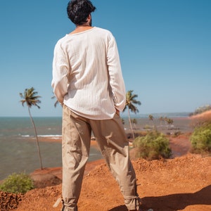 May include: A person wearing a white long-sleeved shirt and beige pants stands on a red dirt cliff overlooking a blue ocean. Palm trees are visible in the background.