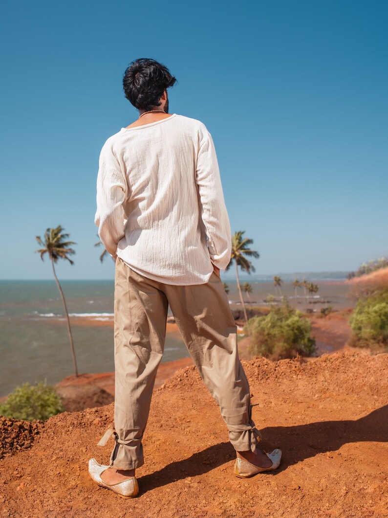 May include: A person wearing a white long-sleeved shirt and beige pants stands on a red dirt cliff overlooking a blue ocean. Palm trees are visible in the background.