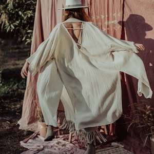 Puede incluir: Una kaftán de lino blanco con la espalda baja y detalles de flecos. La kaftán la lleva puesta una persona con un sombrero blanco y está de pie frente a un fondo rosa.