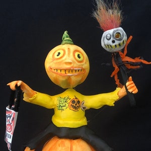 May include: A whimsical Halloween figurine featuring a pumpkin-headed character atop a carved pumpkin. The figure wears a yellow shirt with a pumpkin graphic, black pants, and sneakers. It holds a banner that says "Halloween". A smaller skull figure is held in its other hand.
