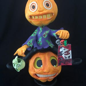 May include: A whimsical Halloween figurine featuring two smiling orange pumpkin heads stacked on top of each other. The top pumpkin wears a purple shirt with green accents and holds a green skull. A Trick or Treat bag is held by the top pumpkin.