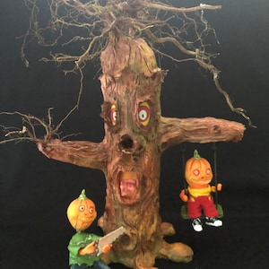 May include: A Halloween-themed tree sculpture with a carved face, arms, and exposed roots. Two pumpkin-headed figures are featured, one on a swing and the other holding a saw. The tree and figures are painted in earthy tones with red and yellow accents.