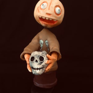 May include: A whimsical Halloween figurine featuring a pumpkin-headed figure with a wide grin, holding a skull. The figure wears a brown shirt with a skeletal design and stands on a brown base. The pumpkin head has a green stem.