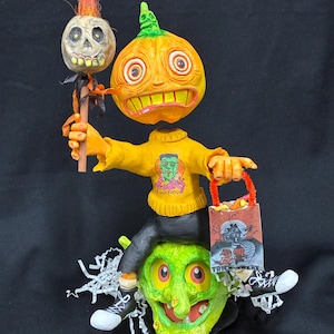 May include: A whimsical Halloween figurine featuring a pumpkin-headed figure wearing a yellow sweater with a Frankenstein graphic, seated on a green witch's head. The pumpkin figure holds a skull on a stick and a trick-or-treat bag. The figurine is decorated with black and white sneakers.