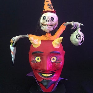 May include: A whimsical Halloween sculpture featuring a red devil head with yellow eyes and horns, topped with a skeleton wearing a party hat. The skeleton holds a trick-or-treat bag and a spooky head.