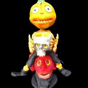 May include: A Halloween decoration featuring a stack of three figures. The top figure is a yellow pumpkin with a green hat, the middle figure is a white skull, and the bottom figure is a red devil with a black beard.