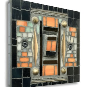 May include: Square mosaic art piece with a geometric design. The mosaic features black, orange, white, and light green tiles, accented with gold-colored metal embellishments and screws. The artwork is mounted on a gray surface.