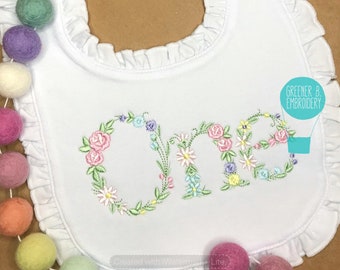 May include: White baby bib with a ruffled edge and embroidered floral design spelling "One".