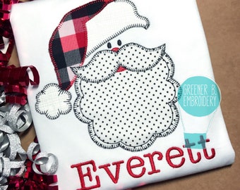 May include: White t-shirt with a red and black plaid Santa applique. The applique has a white beard with black polka dots. The name Everett is embroidered in red below the applique.