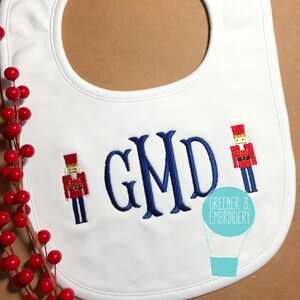 May include: White baby bib with blue embroidered monogram "GMD" and two red and blue embroidered nutcracker figures. The bib has a hot air balloon logo with the text "Greener B. Embroidery".
