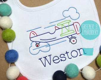 May include: White baby bib with an embroidered airplane design in blue, red, and green. The name "Weston" is embroidered in blue script below the plane. A colorful felt ball garland decorates the left side.