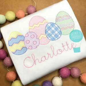 May include: A white t-shirt with a pink, blue, yellow, and green embroidered design of Easter eggs. The name "Charlotte" is embroidered in pink below the eggs. The text "Greener B. Embroidery" is in the top right corner of the image.