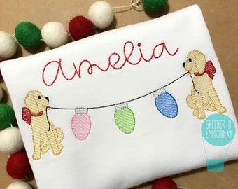 May include: A white embroidered shirt with the name "Amelia" in red thread. Two yellow dogs are pulling a string of colorful Christmas lights.
