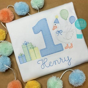 May include: A white onesie with blue and white gingham number one, a white goose with a party hat, and presents. The onesie is personalized with the name "Henry".