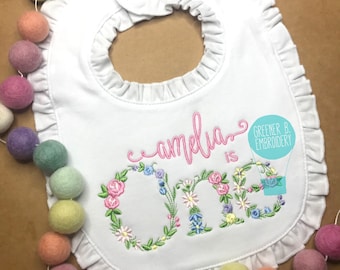 May include: White bib with a pink floral embroidered design that says "amelia is one". The bib has a ruffled edge and is decorated with a small blue hot air balloon with the text "Greener B. Embroidery".
