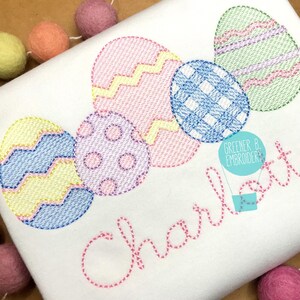 May include: A white t-shirt with embroidered pastel colored Easter eggs and the name "Charlotte" in pink thread. The text "Greener B. Embroidery" is also embroidered in blue thread.