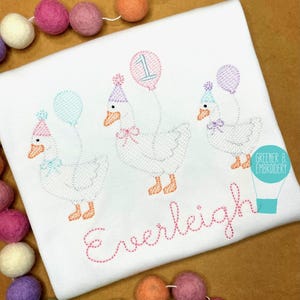 May include: White embroidered shirt featuring three geese wearing party hats and holding balloons. The number "1" is on one balloon. The name "Everleigh" is embroidered in cursive. A garland of colorful felt balls is in the background.