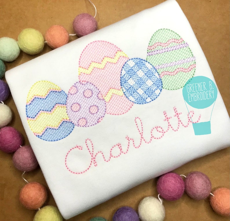 May include: A white t-shirt with a pink, blue, yellow, and green embroidered design of Easter eggs. The name "Charlotte" is embroidered in pink below the eggs. The text "Greener B. Embroidery" is in the top right corner of the image.